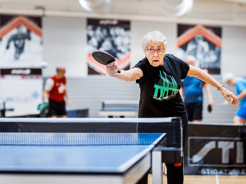 Dam smashar iväg en pingisboll. Hon ser bestämd ut, två personer spelar pingis i bakgrunden.